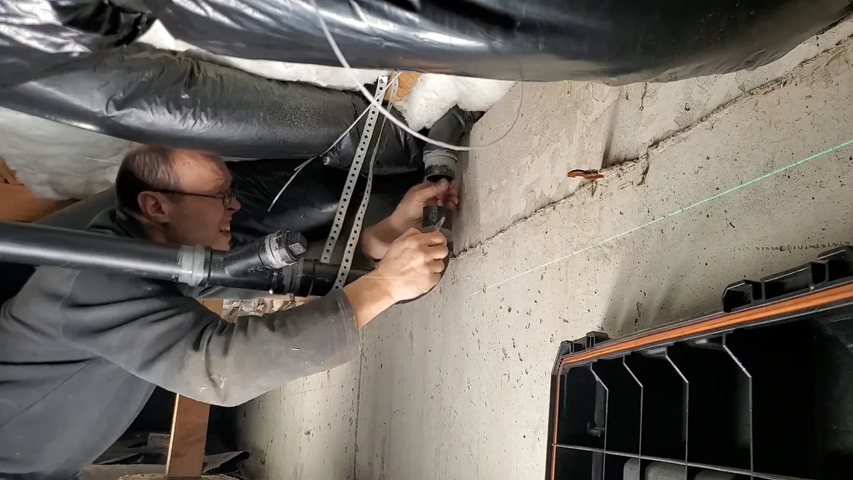 Man in a basement reaching up to pipework under floor joists, with a green laser line across the wall